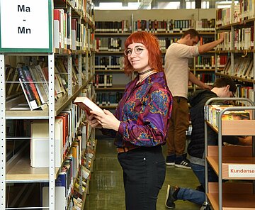 Eine Bibliothekarin steht vor einem Bücherregal und hält ein Buch in der Hand.