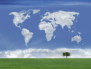 Das Symbolbild zeigt eine Weltkarte aus Wolken über grüner Wiese.