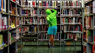 Ein Jugendlicher steht mit neongrüner Jacke und blauer kurzer Hose vor einem vollen Bücherregal. Er ist von hinten zu sehen.