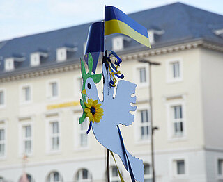 Das Bild steht symbolisch für die Ukrainehilfe. Es zeigt eine weiße Friedenstaube aus Holz mit einer Flagge in den ukrainischen Nationalfarben gelb und blau.