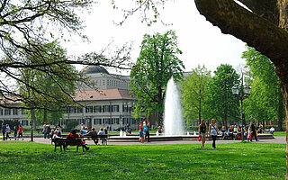 Das Bild zeigt den bevölkerten Friedrichsplatz mit die Rasenfläche, Ruhebänken und der Wasserfontäne. 