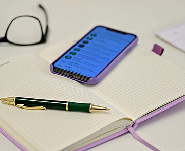 Bild zeigt einen Tisch. Auf diesem liegen ein Notizbuch, ein Smartphone, ein Kuli, eine Brille. Im Smartphone-Display ist ein Kalender zu sehen.