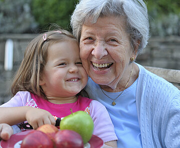 Ältere Frau und Mädchen sitzen an einem Tisch mit einem Teller Äpfel und lächeln in die Kamera