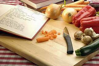 Das Symbolbild zeigt ein Schneidebrett mit Messer, Gemüse und Kochbuch.