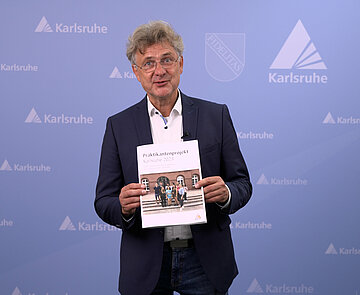 Oberbürgermeister Dr. Frank Mentrup der Stadt Karlsruhe hält die Abschlussarbeit zum Sommerpraktikum 2023 - der Praktikantinnen und Praktikanten aus Karlsruhes Partnerstädten -  in der Hand.
