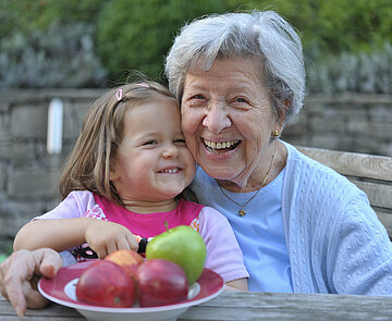 Ältere Frau und Mädchen sitzen an einem Tisch mit einem Teller Äpfel und lächeln in die Kamera