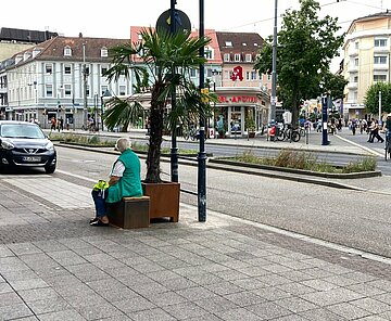 Sitzgelegenheiten und Grünelemente in der Karlstraße: Blick auf die Karl-Apotheke und die Karlstraße. Auf dem Gehweg befindet sich ein Kübel mit einer Palme. Davor sitzt eine Person auf einer Bank.
