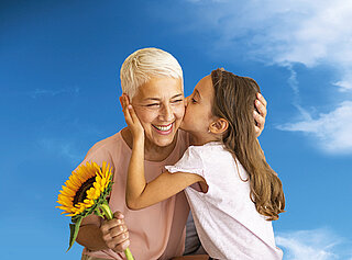 Das Bild zeigt eine Frau mit Sonnenblume in der Hand, die von einem Mädchen umarmt und auf die Wange geküsst wird.