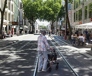 Das Bild steht symbolisch für Menschen mit eingeschränkter Mobilität. Es zeigt eine ältere Dame mit Gehhilfe, die eine Straße in der Karlsruher Innenstadt überquert.