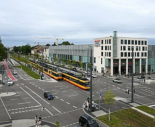 Blick von oben auf die Kreuzung Ettlinger Tor, die begrünten Bahngleise, zwei Bahnen an der Haltestelle und Fußgänger.