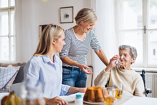 Ältere Frau sitzt mit zwei jüngeren Frauen am Tisch und trinkt ein Glas Wasser.