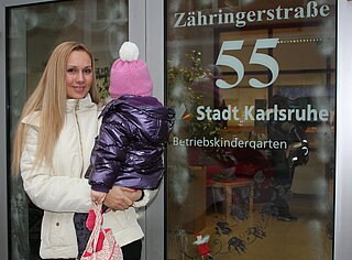Junge Frau mit Kind auf dem Arm steht vor dem Kindergarten der Stadt Karlsruhe