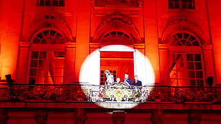 St. Nicolas und die Oberbürgermeister von Karlsruhe, Nancy und Winnyzja stehen auf dem Balkon des Rathauses von Nancy, das rot angestrahlt ist