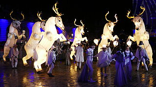Sankt Nikolaus-Umzug mit weiß leuchtenden Rentieren auf den Hinterläufen, die von hell bekleideten Menschen mit Umhang und Hut geführt werden