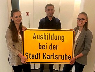 Zwei junge Frauen und ein junger Mann halten ein Schild mit Aufschrift Ausbildung bei der Stadt Karlsruhe.