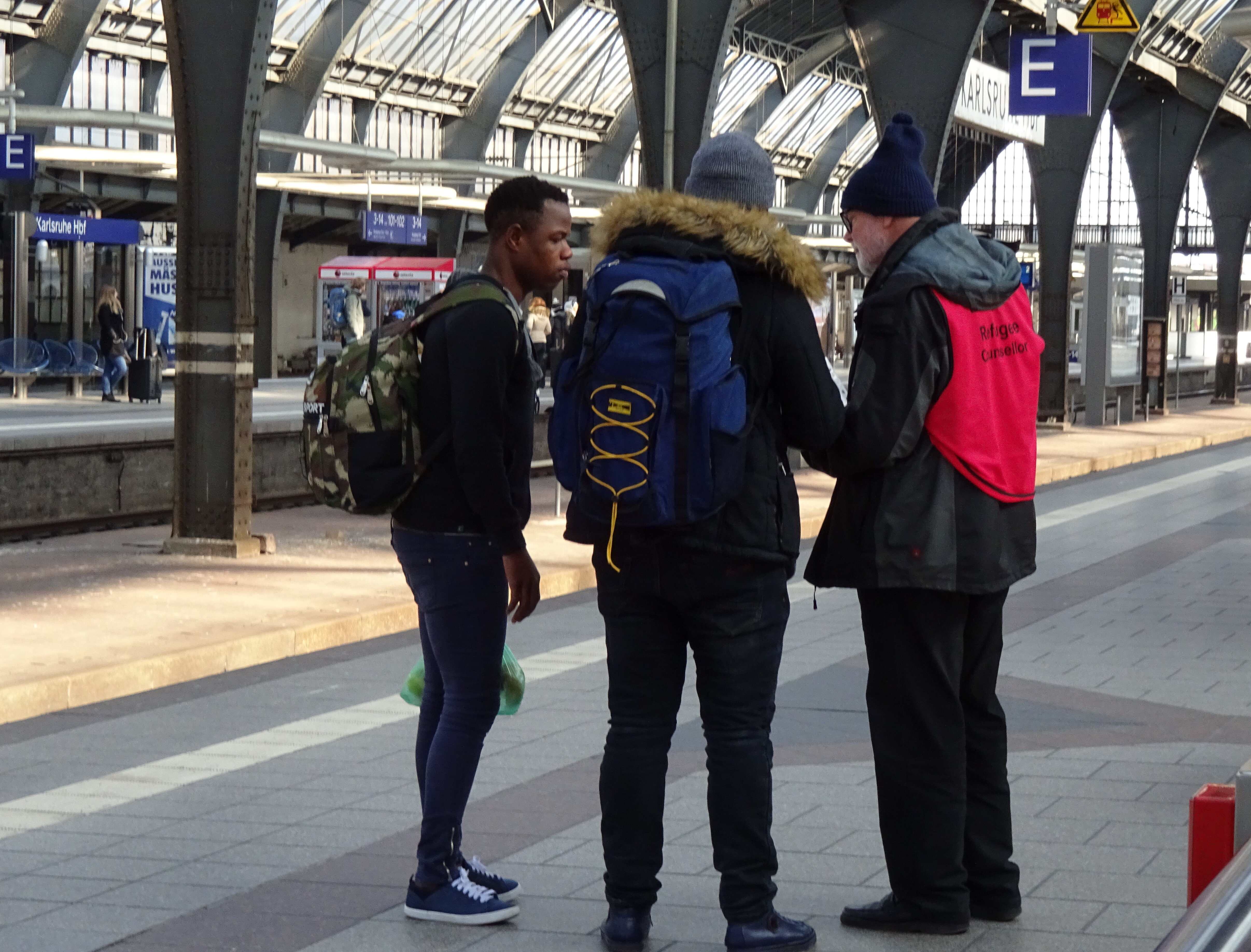 Das Bild steht symbolisch für die Flüchtlingsberatung der Caritas und Diakonie Karlsruhe. Es zeigt drei Personen in einer Gesprächssituation am Karlsruher Hauptbahnhof.