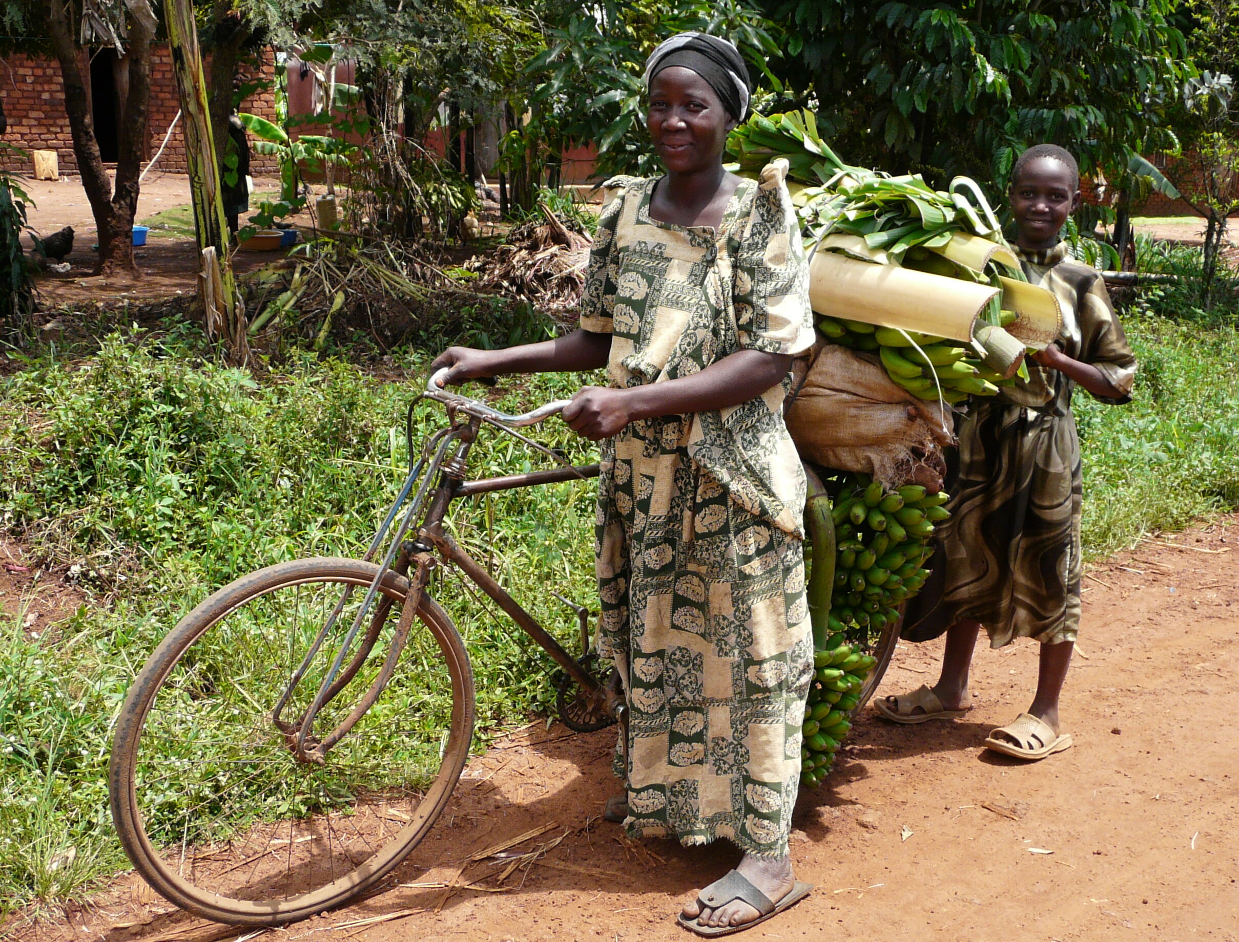 Das Bild zeigt eine Frau mit einem vollgepakcten Fahrrad und ein Kind in Uganda.