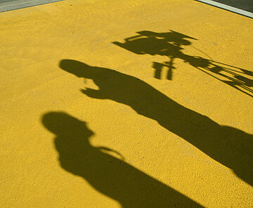 Das Bild zeigt den Schatten von zwei Personen und einer Kamera mit Stativ vor einem gelben Boden.